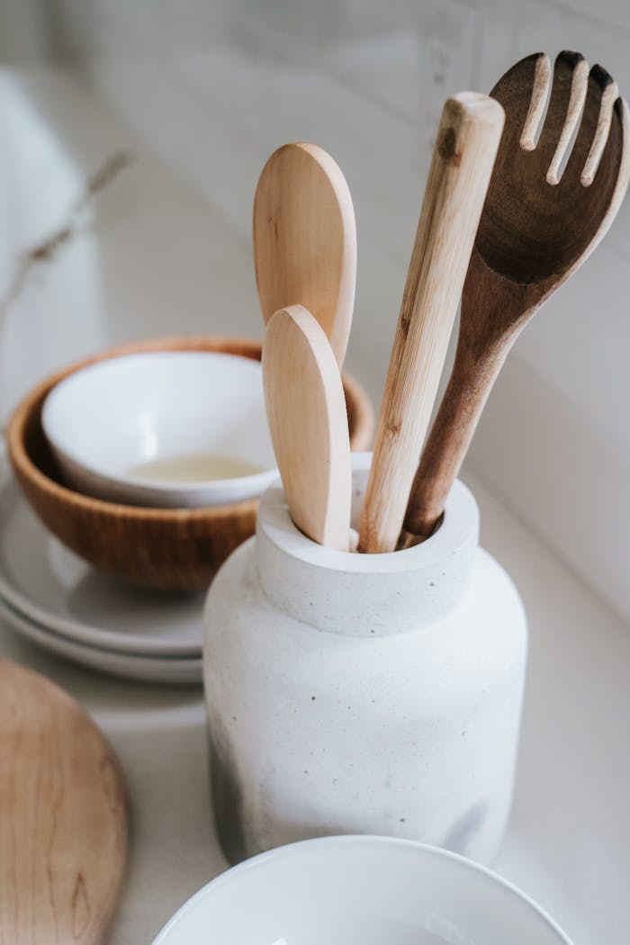 Stylish arrangement of wooden cooking utensils in ceramic jar with bowls on white countertop.